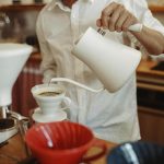 Barista w białej koszuli używa białego czajnika gooseneck do precyzyjnego zalewania kawy w dripperze Hario V60, stojąc przy drewnianym blacie w kawiarni.
