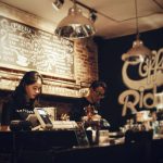 Kawiarnia specialty wpisująca się w czwartą falę kawy – baristka przygotowuje zamówienia w klimatycznym wnętrzu z tablicą menu i industrialnym oświetleniem.