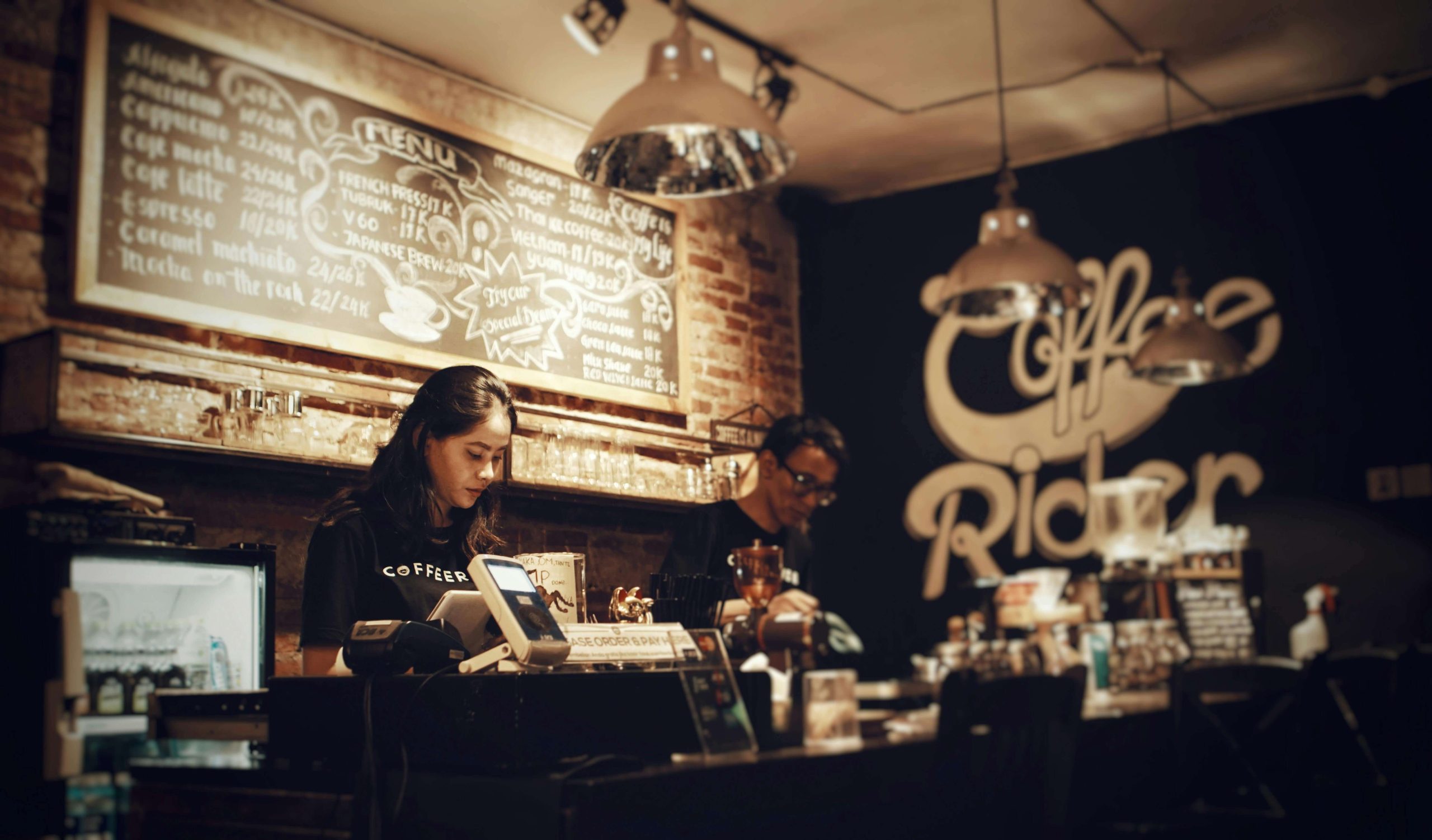 Kawiarnia specialty wpisująca się w czwartą falę kawy – baristka przygotowuje zamówienia w klimatycznym wnętrzu z tablicą menu i industrialnym oświetleniem.