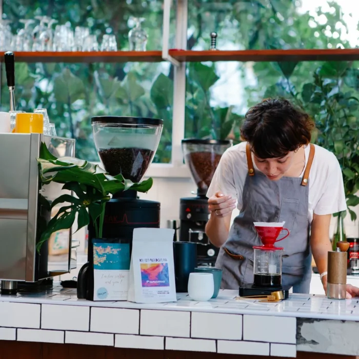 Młoda kobieta-barista w fartuchu przygotowuje kawę rzemieślniczą metodą przelewową (dripper) na wagdze w kawiarni.
